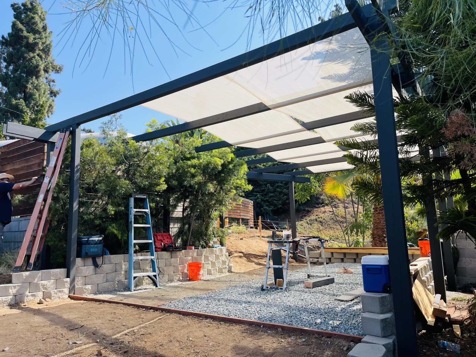 Steel framed shade structure with fabric panels over a gravel patio and block retaining wall
